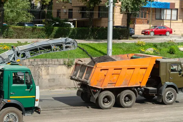 Değirmen makinesi yıpranmış asfalt yüzeyini kaldırır, taşıyıcı enkazı turuncu damperli kamyona yükler, yeşil kamyon yardımcıları, şehir sokakları, yol çalışmaları, yeniden yapılanma, altyapı gelişimi.
