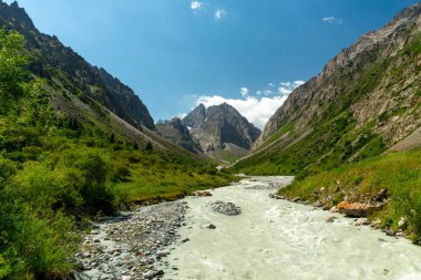 Dağ nehri, sarp kayalıklarla çevrili yemyeşil vadi boyunca akar, yukarıdaki açık gökyüzü, dağcıları ve dağcıları uzak yabanı keşfetmeye ve dağlık alp maceralarını yaşamaya davet eder..
