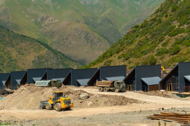 Yeşil tepeler, inşaat araçları, toprak taşıma ekipmanları ile çevrili bir dağda, İskandinav kireçtaşı, bir çerçeveli evler ve stilt evler inşa ediliyor. İnşaat malzemeleri görülebilir durumda.