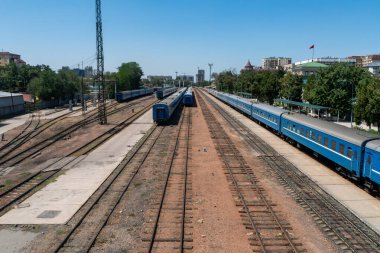Farklı raylara yerleştirilmiş çoklu mavi vagonlar, istasyon binaları, sinyal direkleri, semafor sinyali, katenareri kutupları, katılım anahtarları, yan hatlar ve şehir manzarası, operasyonel demiryolu ortamı.