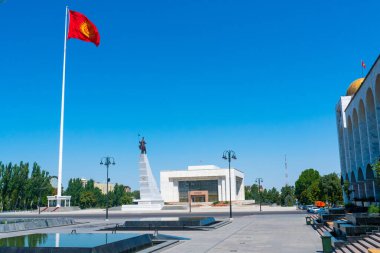 Bişkek 'teki Ala-Too Meydanı' nda Manas Anıtı, Kırgızistan bayrağı, Tarih Müzesi, çeşmeler, açık plaza, ağaçlar ve çevresindeki kentsel manzara, ulusal gurur ve mirası sembolize ediyor..