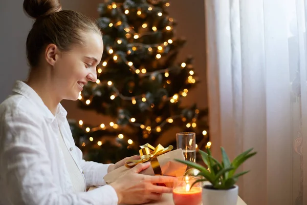 Beyaz gömlekli, gülümseyen güzel bir kadının profil portresi Noel ağacının yanındaki masada oturuyor ve hediye kutusunu elinde tutuyor, çok mutlu oluyor, tatilleri bekliyor..