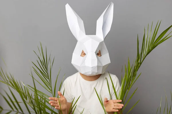 stock image Indoor shot of unrecognizable woman wearing white shirt and rabbit mask posing isolated over gray background, standing among palm branches looking at camera.