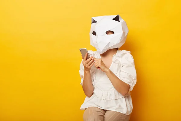 stock image Portrait of anonymous woman blogger wearing dress and paper cat mask holding cell phone, writing post for her blog, using mobile phone, posing isolated over yellow background.