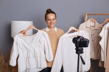 Bun saç stili video ile gülümseyen sevimli bir kadının portresi, tripoyla kamera kullanmak, askılarda kıyafet tutmak, en iyi giyim tarzını seçmek..