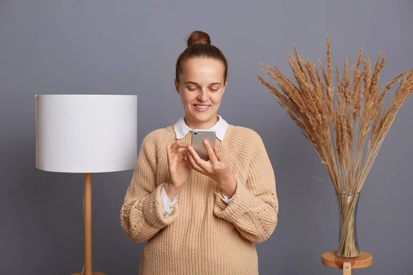 stock image Horizontal shot of smiling joyful woman wearing beige warm sweater standing indoor near lamp in living room, holding mobile phone, checking social networks, reading messages, typing.