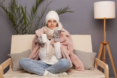 Portrait of winsome sad upset cold woman sitting on the sofa at home drinking hot tea and using smart phone, female wearing winter coat, hat and mittens. clipart