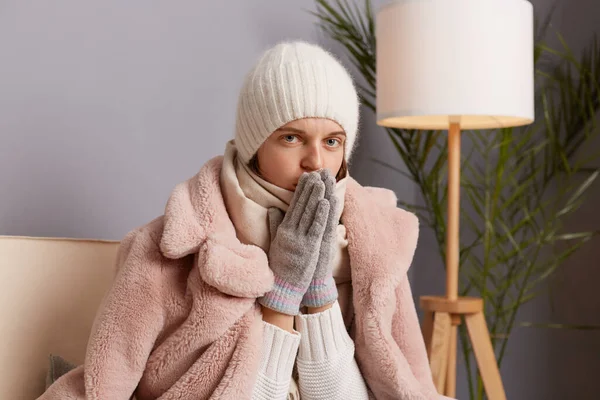 Indoor shot of freeze woman sitting on the sofa at home wearing winter coat, hat and mittens, warms hands in a cold house, looking at camera, have cold.