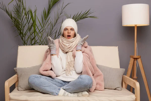 Shocked angry Caucasian woman sitting on the sofa at home, looking at camera with open mouth and big eyes, asking why central heating is turn off, female wearing winter coat, hat and mittens.