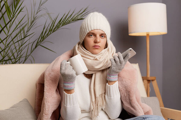 Image of Caucasian attractive woman sitting on the sofa at home wearing winter coat, hat and mittens, holding smart phone in hands, looking at camera, posing with cup in hands, drinking hot tea.