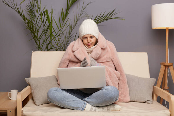 Portrait of Caucasian freezewoman freelancer sitting on the sofa at home wearing hat and mittens, wrapped in coat? working online on laptop and trying to get warm.