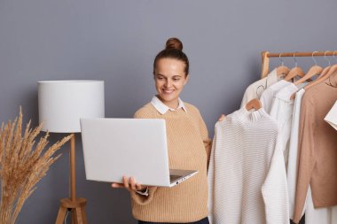 Moda mağazasında askıda giysileri tutan, dizüstü bilgisayarını tutan ve vlog 'unda abonelerine şık kıyafetler gösteren iyimser, mutlu bir kadının portresi..