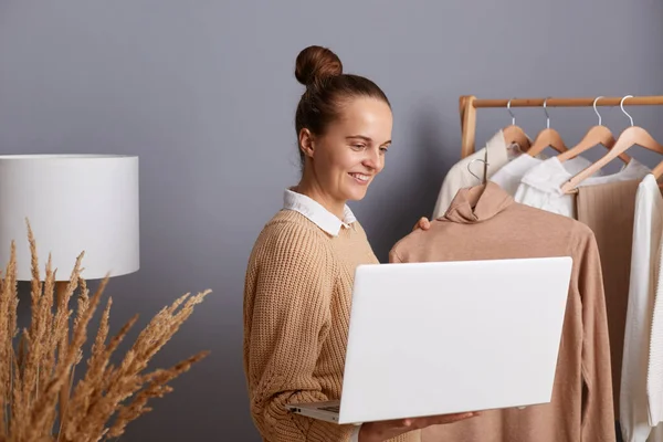 Memnun bir kadın alıcının galeride askıda süveterini tutarken müşterisine Jumper 'ı gösterirken elinde dizüstü bilgisayarla olumlu duygular ifade ederken görüntüsü..
