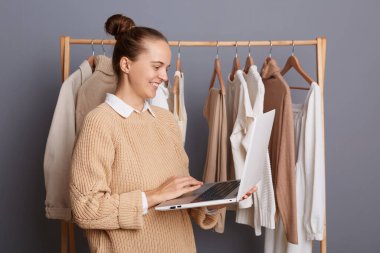 Gülümseyen kadın tasarımcı portresi moda mağazasında elbise askısının yanında duruyor ve dizüstü bilgisayarı tutuyor, internette çalışıyor, yeni kıyafetler için eskizler çiziyor..