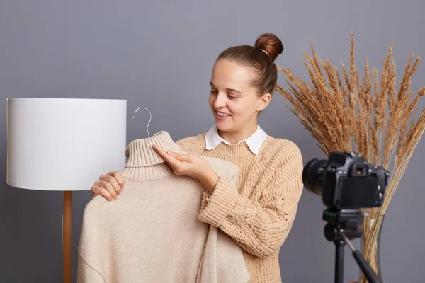 満足した幸せな女性の屋内ショットは ランプとドライフラワーとグレーの壁にベージュのセーターを着て カメラで三脚の前に立って ビデオを記録し 彼女の服を販売 — ストック写真