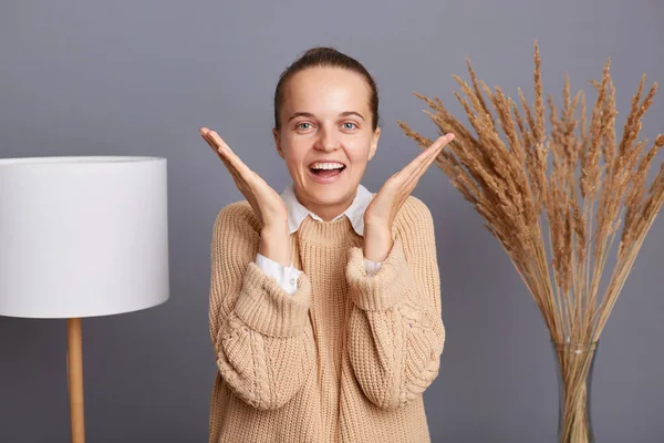 Portrait of extremely happy surprised woman wearing beige sweater standing against gray wall with lamp and dried flowers, raised arms, screaming happily, hearing good news.