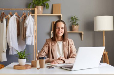 Ceketli, neşeli, genç, beyaz kadın tasarımcının tasviri. İç ofiste yeni çizimlerle çalışıyor. Giysiler rafta asılı, mutlu bir ifadeyle kameraya bakıyor..