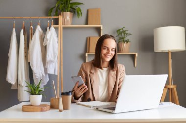 İç mekanda, bej ceketli güzel beyaz bir kadının dizüstü bilgisayarda çalışırken, rafta asılı cep telefonunu tutarken, dizüstü bilgisayara bakarken çekilmiş..