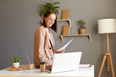 Çekici, koyu renk saçlı, bej ceketli, masada oturan, defter üzerinde çalışan, elinde belgelerle eğlenen beyaz bir kadının yatay görüntüsü..