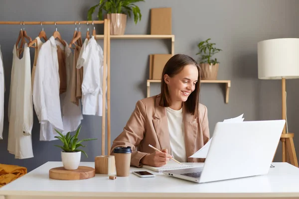 stock image Horizontal shot of beautiful cheerful woman designer in jacket working on computer in living room against clothes hang on shelf, holding papers, drawing sketches.