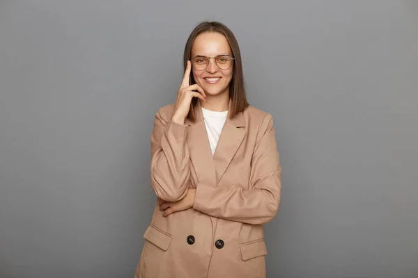 Portrait of self confident successful young adult businesswoman wearing beige jacket, looking at camera with positive expression, posing isolated over gray background.