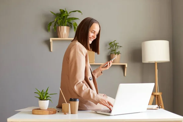 Indoor shot of young adult adorable happy businesswoman wearing jacket working on home office and using laptop and mobile phone, being in good mood at work.