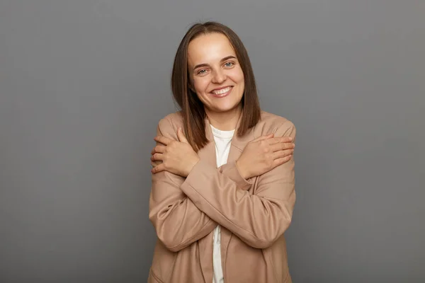 I love myself. Pleased satisfied female wearing beige jacket standing isolated over gray background, hugging herself and smiling, feeling comfortable and fulfilled, narcissistic egoistic person.