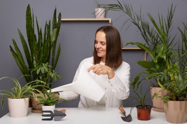 Gülümseyen kahverengi saçlı kadın çiçekçinin portresi çiçekçide çiçekleri sulayan beyaz gömlek giyen, olumlu duygularını ifade eden, mutlu bir şekilde gülümseyen..