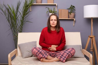 Hasta, kahverengi saçlı, kırmızı kazaklı, kareli pantolonlu, sağlıksız görünüşlü, karnını tutan, güçlü bir karın ağrısı çeken, öksürük odasında oturan hasta bir kadının kapalı çekimleri..