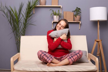 Kırmızı kazak ve pijama giyen, yastığı kucaklayan mutlu, genç bir kadının görüntüsü. Keyfi yerinde, hafta sonları dinleniyor, dinleniyor, evdeki kanepede oturuyor..