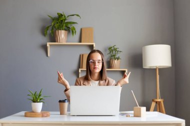 Sakin bir iş kadını meditasyon için ara veriyor, iş yerinde yoga yapıyor, düşünceli çekici bir kadın işçi zen hissediyor, stresten uzak ve uyum içinde çalışıyor, dizüstü bilgisayarın önünde oturuyor..