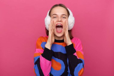 Kış kürkü kulaklık ve kalın süveter giyen, pembe arka planda izole edilmiş, yüksek sesle bağıran, anons yapan, avuç içlerini açık tutan,.