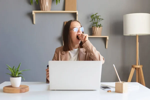 Attractive beautiful young Caucasian female feel tired and overworked, woman in glasses work remotely from home or sitting at table in office in front of laptop, yawning and covering mouth.