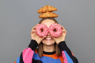 Kapalı alanda renkli kazak giyen, eğlenen, gözlerini donutla kapayan, kafasında kruvasanlarla gri arka planda dikilen neşeli bir kadın resmi..