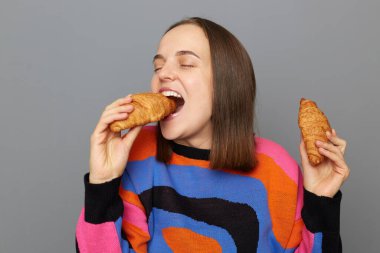 Kapalı alanda, gri arka planda yalnız poz veren, kruvasan yiyen, aç hisseden, şekerli tatlı yemeyi seven, mutlu, beyaz kadın pozu..