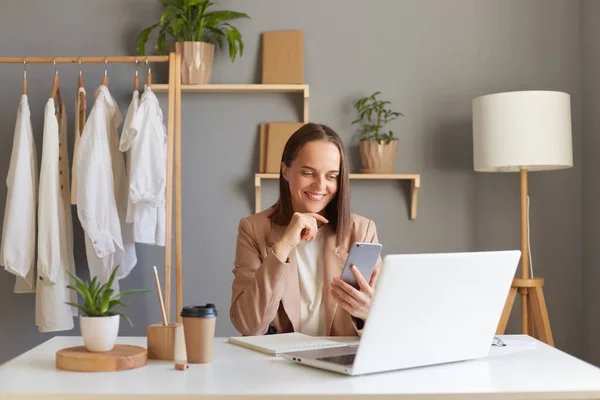 stock image Indoor shot of smiling attractive brown-haired woman wearing beige jacket sitting at her workplace in front of laptop and using smartphone, expressing positive emotions.