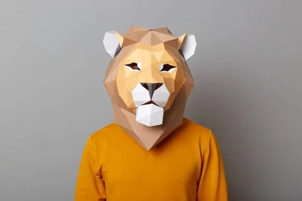 Indoor shot of man wearing orange sweater and lion paper mask standing isolated over gray background, looking at camera, guy in animal costume.