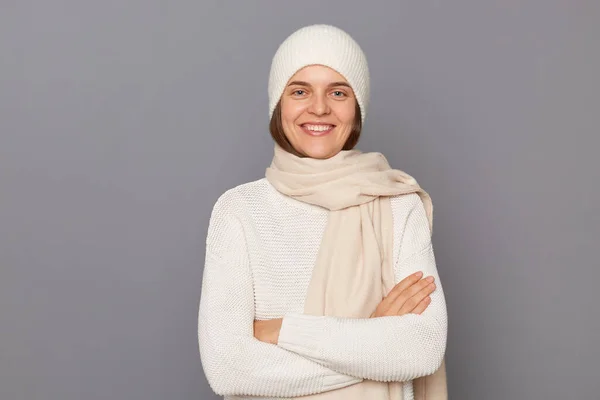 Horizontal shot of smiling delighted satisfied woman wearing white sweater, scarf and cap standing isolated over gray background, keeps hands crossed, looking at camera.