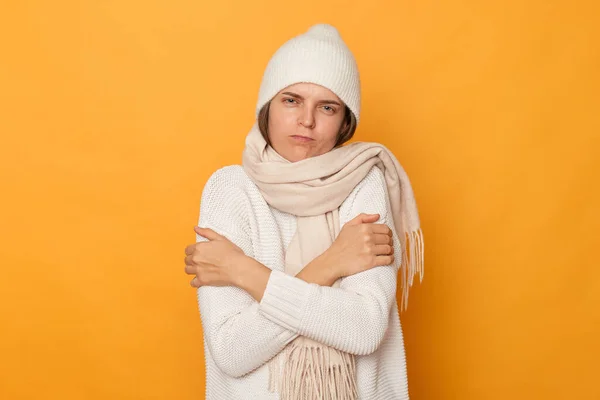 Portrait of sick freeze woman wearing white sweater, scarf and cap standing isolated over yellow background, hugging herself, trying to get warm, expressing sadness, feels cold.