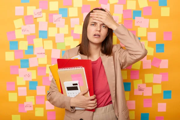 Portrait of frustrated young adult woman having terrible headache or high temperature, studies hard, holds diary with papers, posing against yellow wall with colorful sticky post notes.