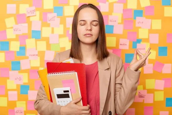 Indoor shot of calm relaxed young adult woman holding papers and calculator, trying to calm down after working or studying, posing against yellow wall with colorful sticky post notes.