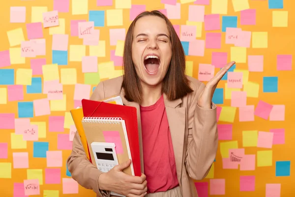 Horizontal shot of brown-haired Caucasian woman manager holding paper documents, isolated over yellow wall covered with stickers, keeps eyes closed, raised hand, screaming.