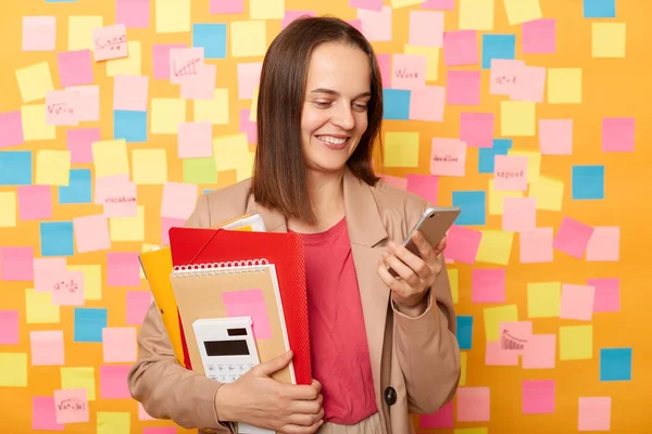 Portrait of attractive positive woman holding paper folders in hands and using cell phone, chatting with friends while having break, posing against yellow wall with stickers for leaving reminder.
