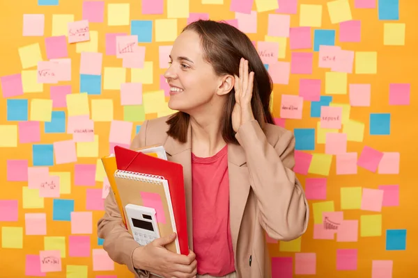 stock image Indoor shot of smiling young adult woman holding papers, posing against yellow wall with colorful stickers, tries to overhear someone, holds hand near ear, tries to understand words.