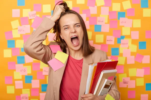 Photo of crazy tired young adult woman holding papers, posing against yellow wall with colorful stickers, shouting very loud, being exhausted, feels fatigue from work or studying, rips hair on head.