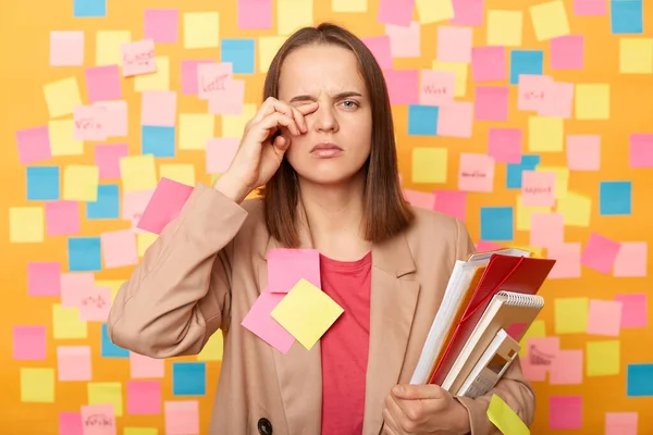 Image of tired stressed young adult woman holding papers, posing against yellow wall with colorful stickers, rubbing her eye, feels pain, looking at camera with pout lips.