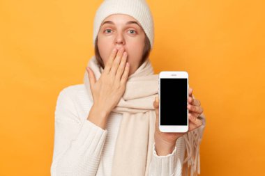 Şaşırmış bir kadın, sıcak süveter, kep ve eşarp içinde duruyor, cep telefonunu boş siyah çizgili gösteriyor, ağzı palmiye, kopya alanı ile kaplıyor, sarı arka planda izole edilmiş..