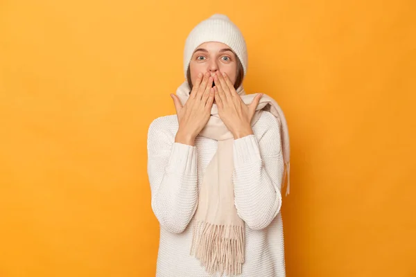 Amazed woman wearing white sweater, cap and scarf standing isolated over yellow background, covering mouth with surprise, being stunned to hear unexpected news, reacts on something.