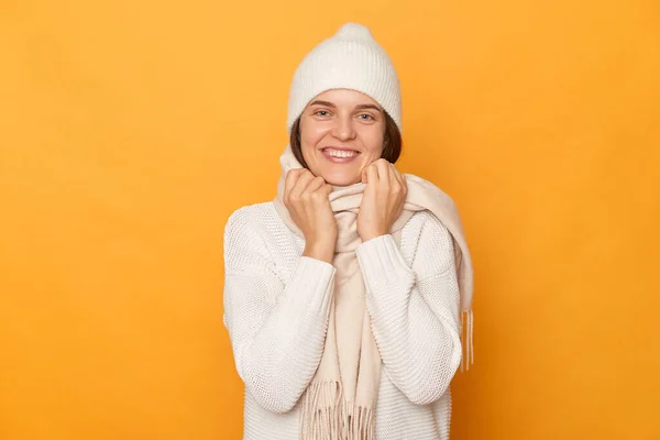 照片中 一个积极快乐的女人穿着白色的连衣裙 帽子和围巾 手挽着领子 面带微笑地看着相机 靠着黄色的墙壁摆姿势 — 图库照片