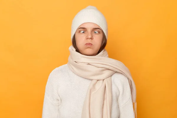Funny hilarious woman wearing white sweater, cap and scarf, crosses eyes, makes funny grimace, pretends to be little fool, plays with kids, being childish, standing isolated over yellow background.